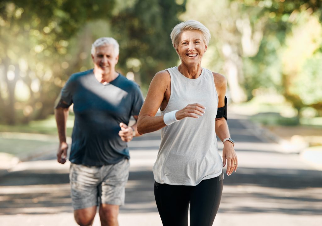 Senior couple wearing sports attire smiling and jogging in park. Lots of trees on the background.