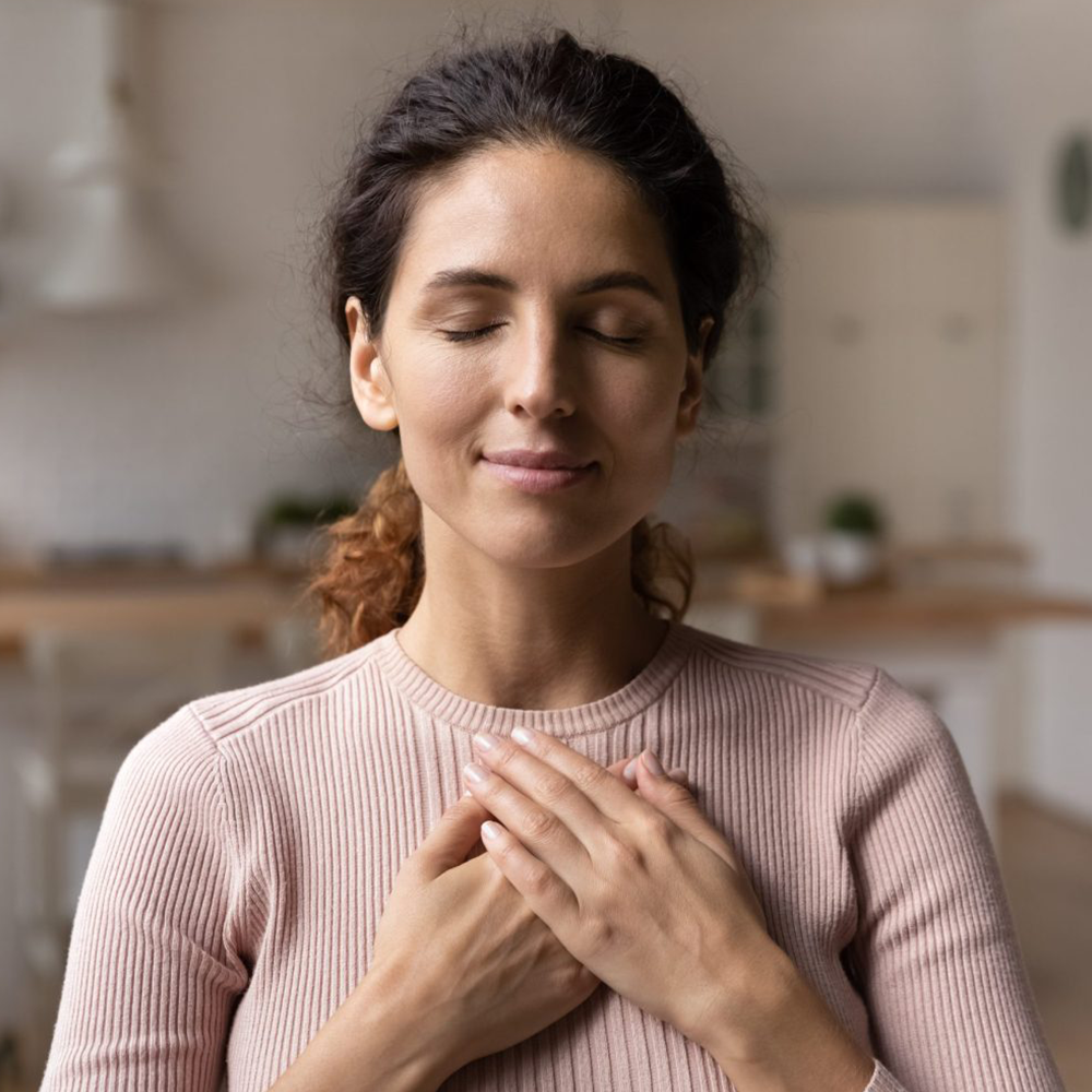 Woman wearing peach shirt with eyes closed and both hands on her chest, smiling and relaxing.